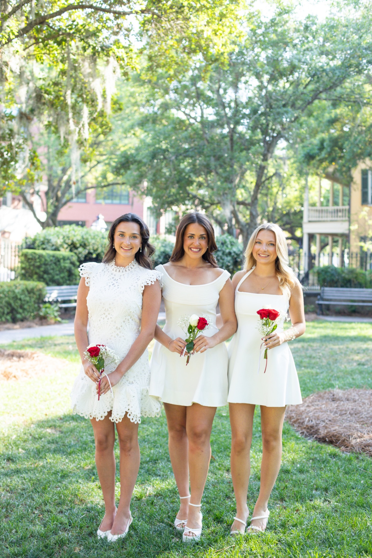 Three friends in white dresses holding red roses in a Charleston park