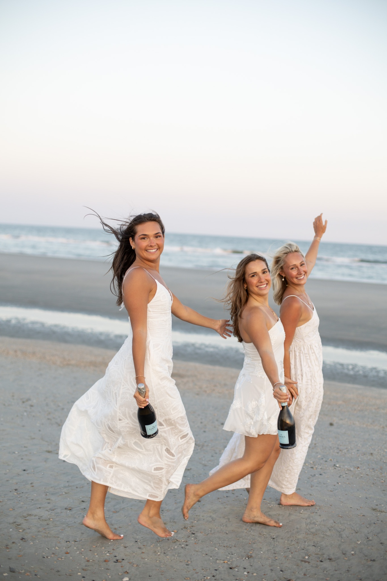 Three friends in white dresses celebrating with champagne on the beach at sunset